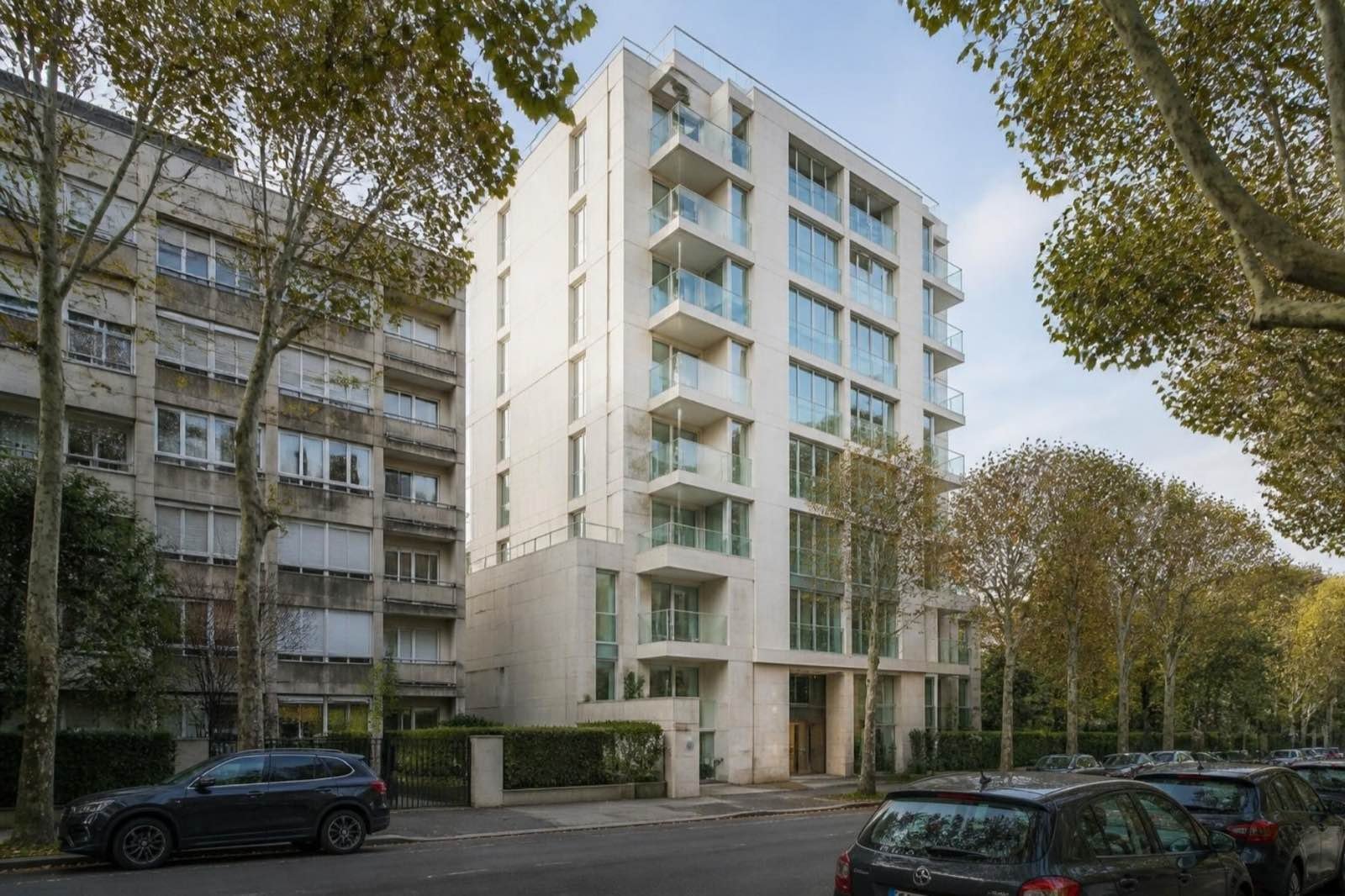 Ağaçlı bir sokakta cam balkonlu, yeni tamamlanmış orta yükseklikte bir apartman; yanında daha eski bir bina